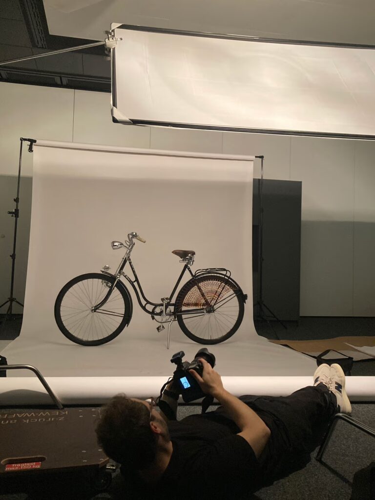 Fotograf liegt auf dem Boden vor Setup mit historischem Damen-Fahrrad vor weißem Hintergrund. Ein Foto ist auf dem Kamera-Display sichtbar.