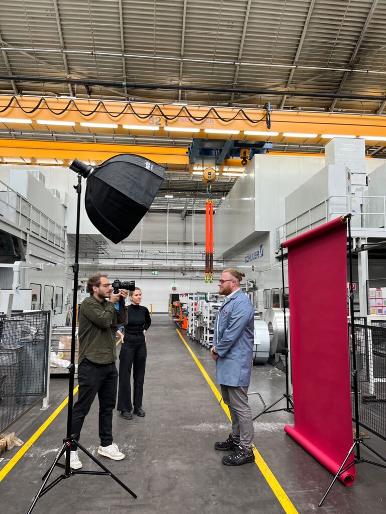 "Behind-the-scenes"-Foto zeigt Shooting-Setup in einer Produktionshalle. Ein Mann in grauem Arbeitskittel wird vor roter Wand fotografiert.