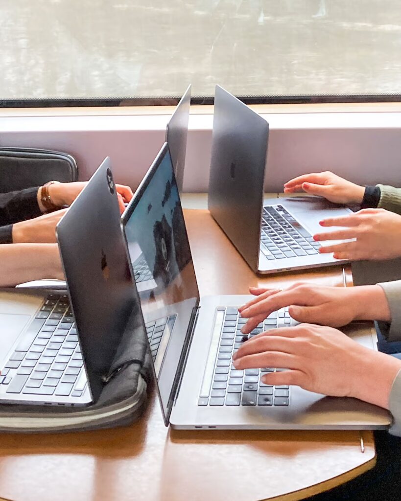 Vier Laptops stehen sich gegenüber auf einem Tisch im Zugabteil, tippende Hände von vier bup-Mitarbeitenden sind zu sehen.