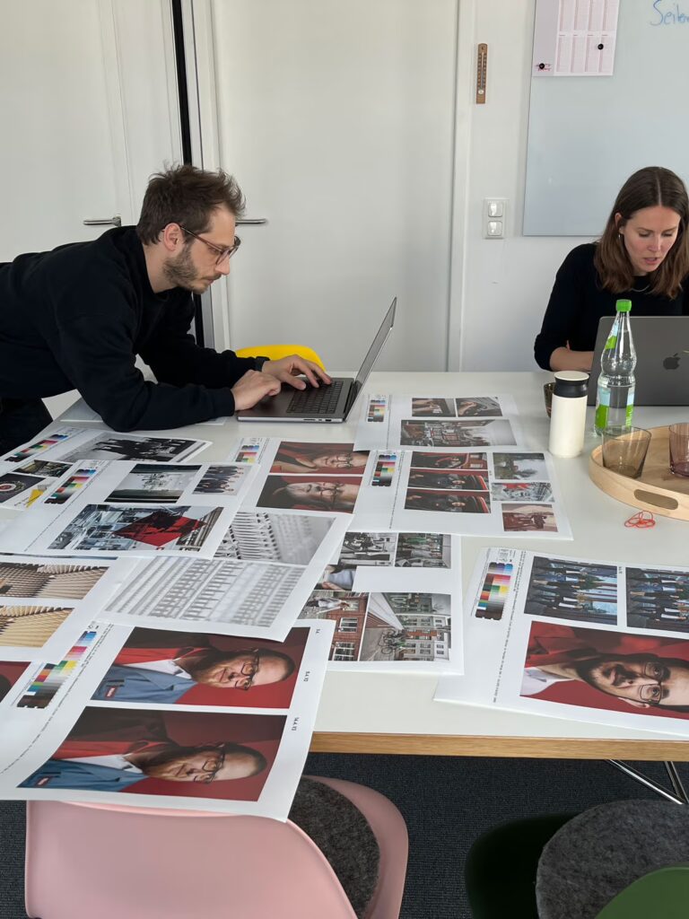 Zwei Mitarbeitende von bup arbeiten an ihren Laptops an einem großen Tisch. Darauf liegen viele Andrucke von Fotos für das Miele Buch.