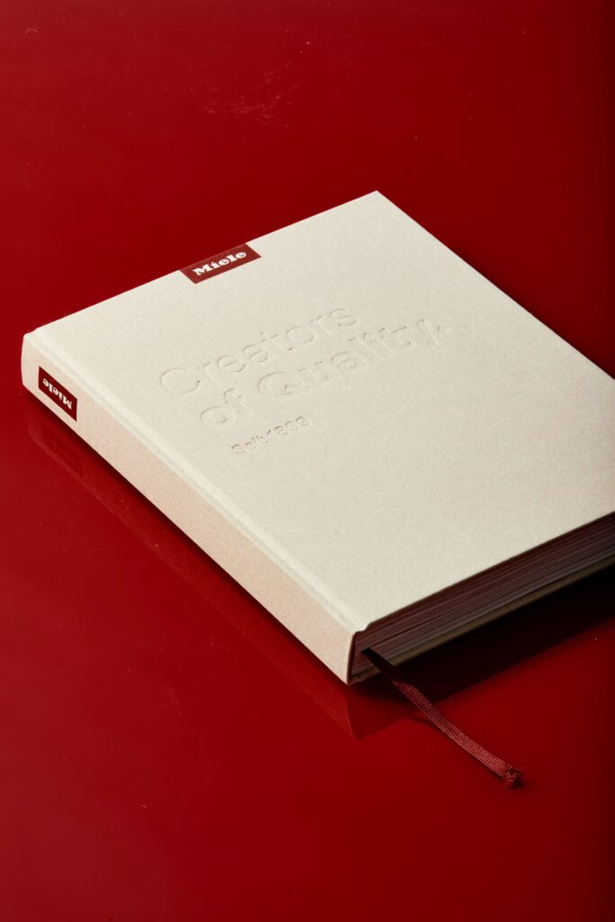 Geschlossenes Miele-Buch liegt auf rotem Untergrund mit leichter Spiegelung.