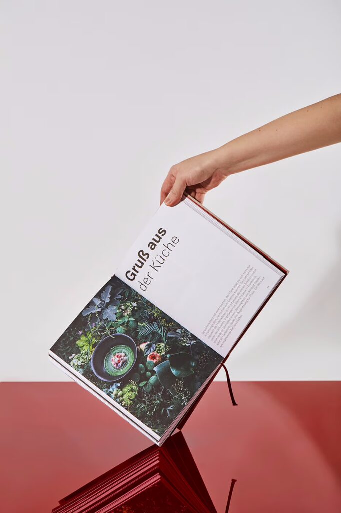 Hand hält "hängendes" Miele-Buch an der rechten oberen Ecke fest. Die aufgeklappte Seite zeigt ein Foto und die Headline "Gruß aus der Küche".
