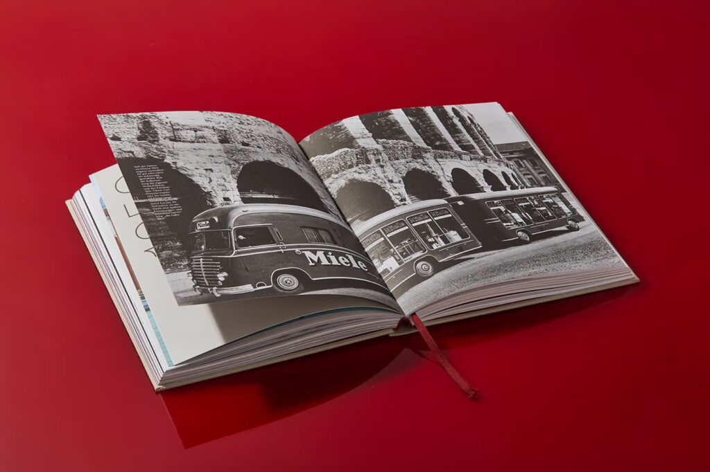 Das Miele-Buch liegt aufgeklappt auf rotem Hintergrund. Es zeigt ein Schwarz-Weiß-Foto eines Miele Ausstellungs-Busses mit Anhänger.