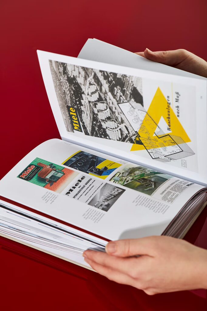 Das Miele Buch liegt aufgeklappt auf rotem Hintergrund. Zwei Hände halten es und blättern darin.