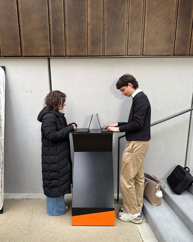 Eine Frau und ein Mann stehen sich gegenüber an einem Pult und arbeiten am Laptop.