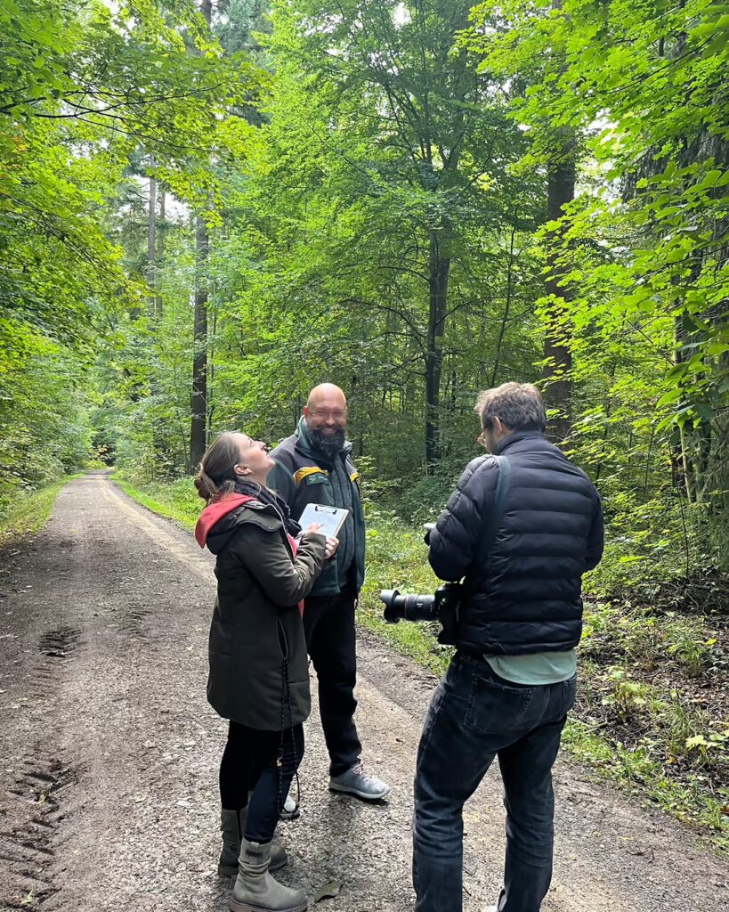 Zwei Personen mit Klemmbrett im Gespräch auf einem Waldweg, daneben ein Fotograf mit Teleobjektiv.