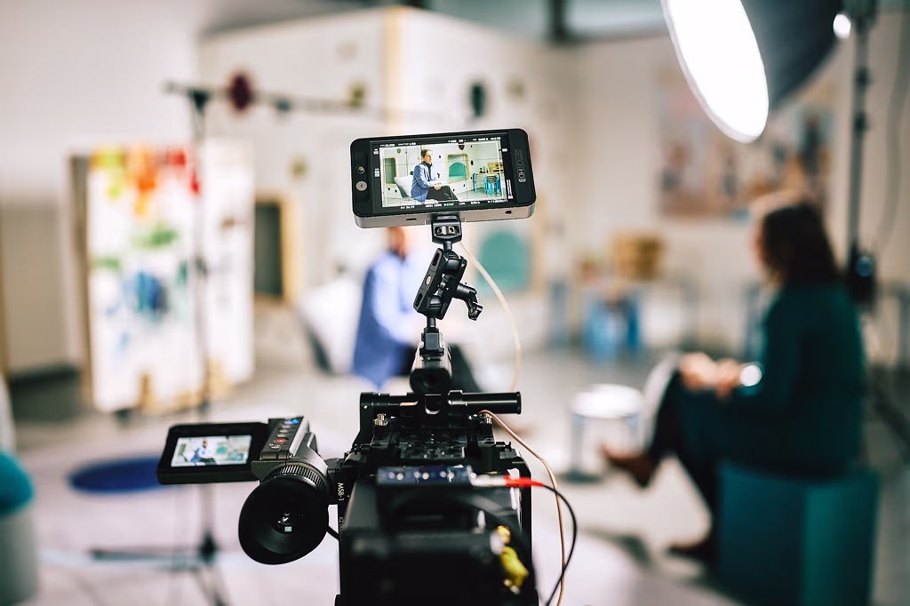"Behind-the-scenes"-Aufnahme zeigt eine Filmkamera im Vordergrund, eine Gesprächssituation unscharf im Hintergrund.