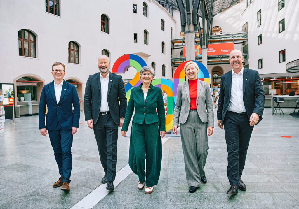 Zwei Frauen und drei Männer gehen auf die Kamera zu und lachen. Im Hintergrund das Jubiläumslogo und ein Filialenfoyer.